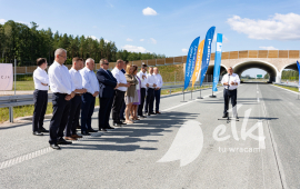 Minister infrastuktury przemawia na drodze Via Baltica z boku stoją oficjalni goście. W tle most i drzewa